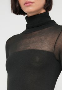 Γυναίκα που φοράει μαύρο διαφανές τοπ με ψηλό γιακά και στερεή μαύρη επιφάνεια που καλύπτει το στήθος, κομμένο στο ύψος της γνάθου.