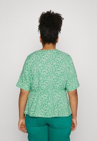 Blusa floral verde con mangas cortas y cintura fruncida, decorada con flores blancas sobre un fondo verde claro.