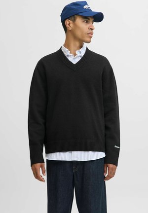 Mand iført en sort V-hals trøje over en hvid skjorte med striber, mørke jeans og en blå baseballkasket, kigger til højre.