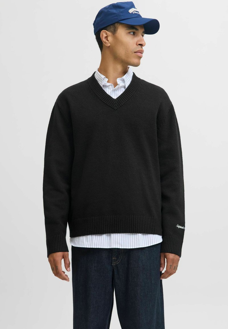 Mężczyzna ubrany w czarny sweter V-neck na białej koszuli w paski z guzikami, ciemne dżinsy i niebieską czapkę z daszkiem, patrzący w prawo.