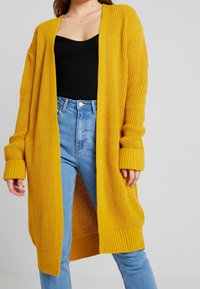 Cárdigan de punto en un amarillo brillante, con mangas largas y un diseño abierto al frente. Textura acanalada en el cuerpo y los puños, combinado con unos vaqueros de tiro alto en azul.