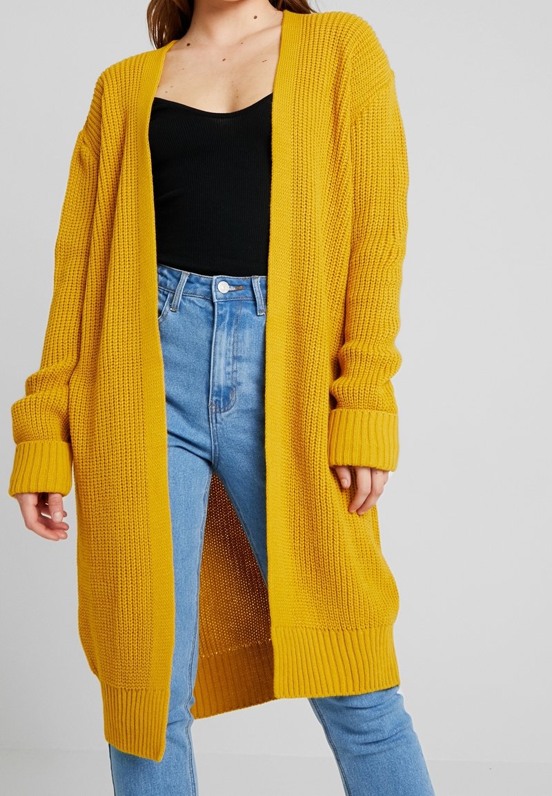 Cárdigan de punto en un amarillo brillante, con mangas largas y un diseño abierto al frente. Textura acanalada en el cuerpo y los puños, combinado con unos vaqueros de tiro alto en azul.