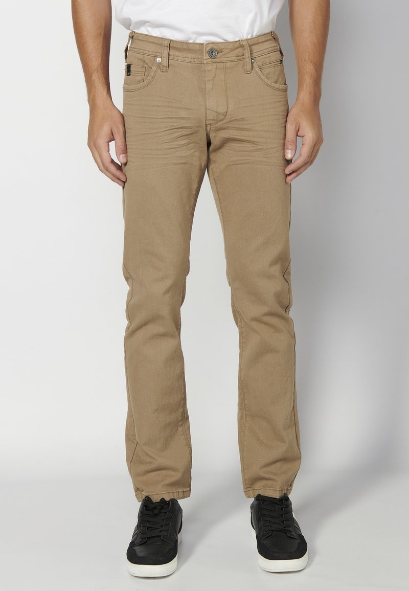 Pantalones de mezclilla en color caqui con un diseño de pierna recta, que presentan un cierre de botón, cinco bolsillos y una textura sutil. Llevados con zapatillas negras.