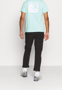 Světlá mátová tričko s grafickým potiskem na zádech, doplněné černými kalhotami a vícebarevnými sportovními botami. Volný střih, ležérní design.