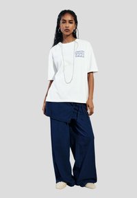 T-shirt blanc avec un graphique bleu clair et un pantalon navy à coupe ample, tous deux en tissu doux. Le modèle porte des chaussures à enfiler plates et texturées.