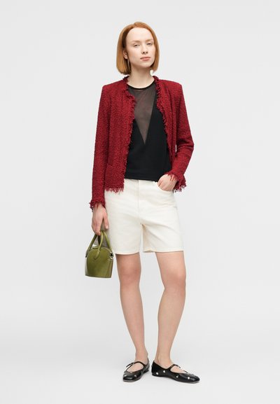 Κόκκινο υφασμάτινο σακάκι με περιθώρια από κρόσσια, μαύρο διαφανές τοπ, λευκό σορτς, μαύρα πάνινα παπούτσια και μια μικρή πράσινη τσάντα. Απλό, μοντέρνο ντύσιμο.