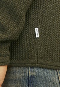 Pulover texturat de culoare verde închis cu tiv striat și model țesut. Eticheta de pe lateral spune "JACK & JONES." Denim albastru vizibil dedesubt.