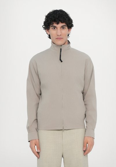 Veste beige clair avec un col haut, fermeture éclair à l'avant et manches longues. Texture lisse, conçue pour un ajustement élégant.