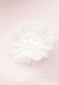 Ροζ ύφασμα με άσπρη βολάν από τούλι και κεντημένο κυκλικό λογότυπο που απεικονίζει τρεις ρίγες και λεπτομέρεια με σχεδιασμό φύλλου.