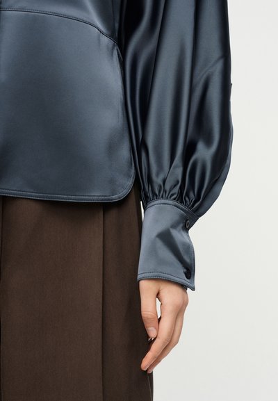 Δεξί χέρι που φοράει μπλούζα από ναυτικό σατέν με μακριά μανίκια, σούρα στον καρπό και μαύρο κουμπί, σε συνδυασμό με σκουρό καφέ φούστα με υφή.