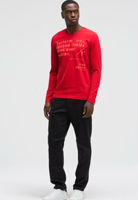 Red long-sleeve shirt with graphic text in gold, paired with black pants and white sneakers. Sleek fit and smooth fabric texture.