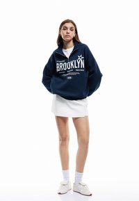 Sudadera con cremallera de color azul marino con gráfico de "BROOKLYN", combinada con una falda blanca y zapatos blancos. Tejido de algodón, corte relajado y mangas largas.