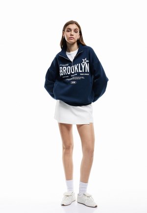 Granatowa bluza z zamkiem z grafiką "BROOKLYN", w połączeniu z białą spódnicą i białymi butami. Wykonana z bawełny, luźny krój i długie rękawy.