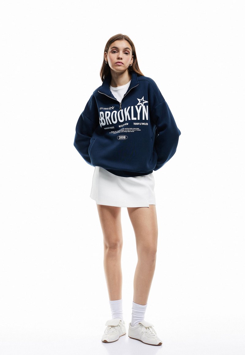 Sudadera con cremallera de color azul marino con gráfico de "BROOKLYN", combinada con una falda blanca y zapatos blancos. Tejido de algodón, corte relajado y mangas largas.