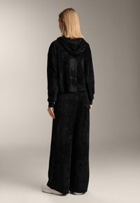 Čierna skrátená mikina s kapucňou, vyrobená z mäkkej látky, v kombinácii s voľnými čiernymi nohavicami. Model je otočený chrbtom, čo ukazuje uvoľnený strih.