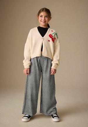 Gestrickte cremefarbene Strickjacke mit roter Blumenstickerei, kombiniert mit schwarz-weiß karierten, weit geschnittenen Hosen und schwarzen Turnschuhen.