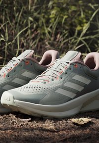 Scarpe sportive verde chiaro in materiale a rete. Le caratteristiche includono dettagli rosa, strisce bianche e un design della suola testurizzato.