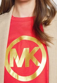 Woman wearing a red shirt with a large gold "MK" logo partially covered by a beige jacket, with long brown hair visible.