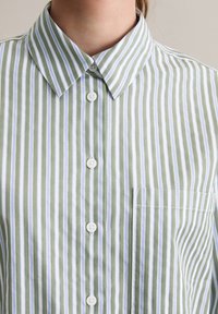 Camicia a righe verde e bianca con colletto, caratterizzata da strisce verticali, bottoni bianchi e una tasca sul petto sinistro. Tessuto liscio e leggero.