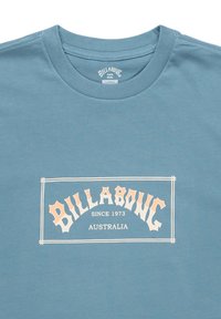 Modrá bavlnená tričko s krátkymi rukávmi, s bielym a oranžovým grafickým textom s nápisom "BILLABONG SINCE 1973 AUSTRALIA" umiestneným na prednej strane.