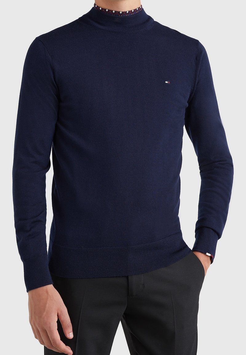 Pull en maille bleu marine avec un col montant, des poignets et un ourlet côtelés. Présente un col intérieur à motifs et un logo discret sur la poitrine.