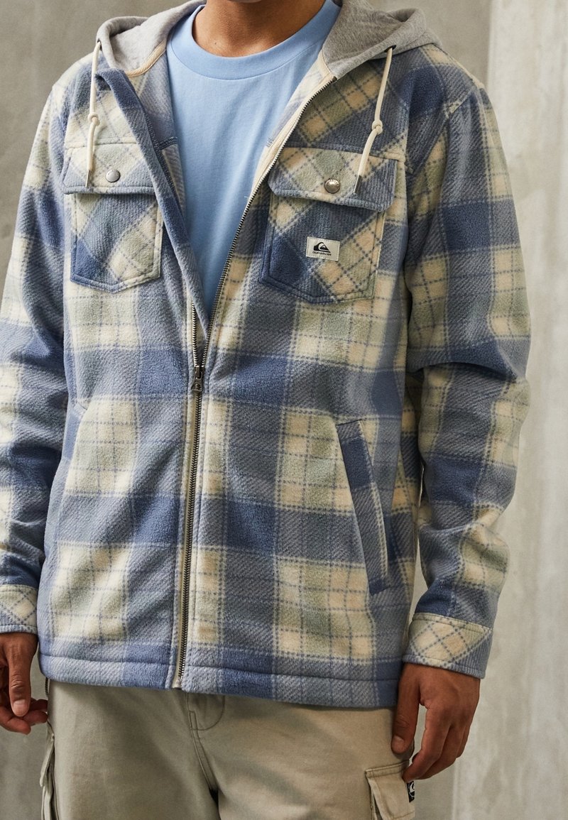 Homme portant une veste polaire zippée à carreaux bleus et beiges avec capuche, par-dessus un t-shirt bleu clair et un pantalon cargo beige.