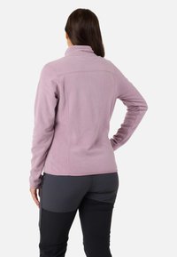 Vaaleanlila fleece-takki, jossa on korkea kaulus ja sileä pinta, raglanhihat ja istuva muotoilu. Mustat housut näkyvät alareunassa.