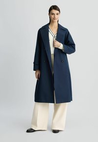 Trench coat doppiopetto blu navy in tessuto impermeabile, presenta bottoni grandi, colletto ampio e vestibilità morbida, indossato sopra un completo crema.