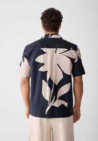 Hombre con pelo corto y rizado, vistiendo una camisa azul marino con un gran estampado floral beige y pantalones beige, visto desde la espalda sobre un fondo liso.