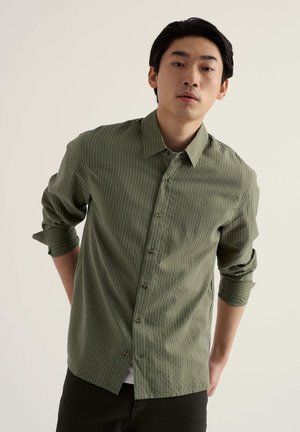 Mladý muž s čiernymi vlasmi, oblečený v zelenú textúrovanú košeľu na gombíky s vyhrnutými rukávmi, stojaci pred jednoduchým svetlým pozadím.