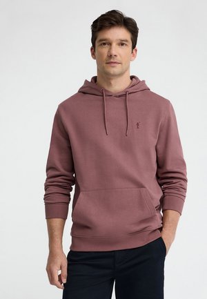 Mężczyzna w fioletowej bluzie z kapturem i kieszenią kangurka oraz ciemnych spodniach, stoi z jedną ręką w kieszeni na jednolitym tle.