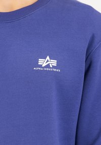 Blå sweatshirt lavet af bomuld med rund halsudskæring. Har et hvidt logo print med et abstrakt design på brystet. Glat tekstur.