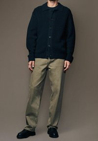 Cardigan en tricot côtelé bleu marine avec col ouvert, fermeture à boutons, associé à un pantalon vert olive clair et des chaussures noires. Design simple et fonctionnel.