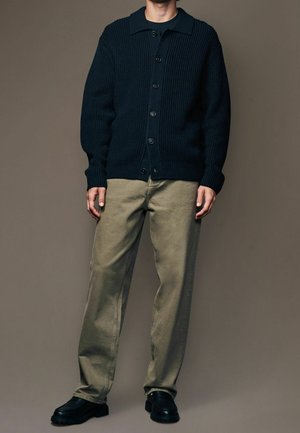 Marineblaues, gerippter Strickcardigan mit einem Umlegekragen und Knopfverschluss, kombiniert mit helloliven Hosen und schwarzen Schuhen. Einfaches, funktionales Design.