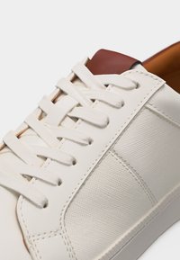 Baskets en cuir blanc avec panneau latéral texturé, lacets blancs et détail en cuir marron sur la languette, montrées en gros plan.