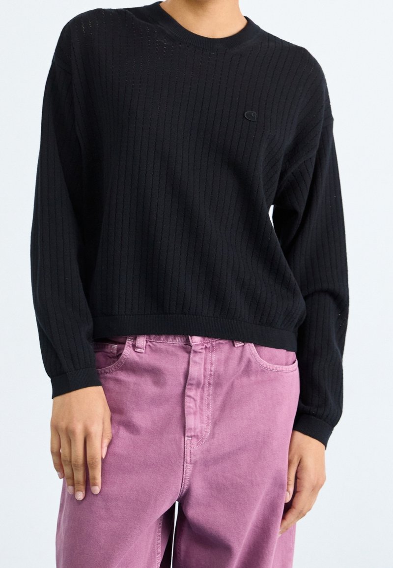 Personne portant un pull noir à côtes manches longues et un pantalon rose taille haute avec poches, debout devant un fond uni.