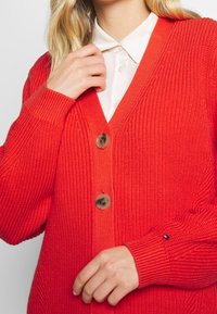 Cardigan de punto rojo con cuello en V, dos botones, puños acanalados y un sutil acento con el logo en la manga.