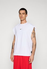 Man with short dark hair wearing a sleeveless white Nike shirt and red shorts, showing tattoos on both arms against a plain background.
