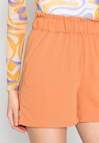 Oranje shorts met een gestructureerde stof, voorzien van een gerimpelde tailleband en zijzakken, gecombineerd met een kleurrijke top met lange mouwen.