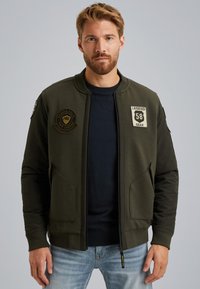 Veste bomber vert olive avec un haut texturé, fermeture éclair, poignets côtelés, deux poches avant et des patchs brodés sur la poitrine.