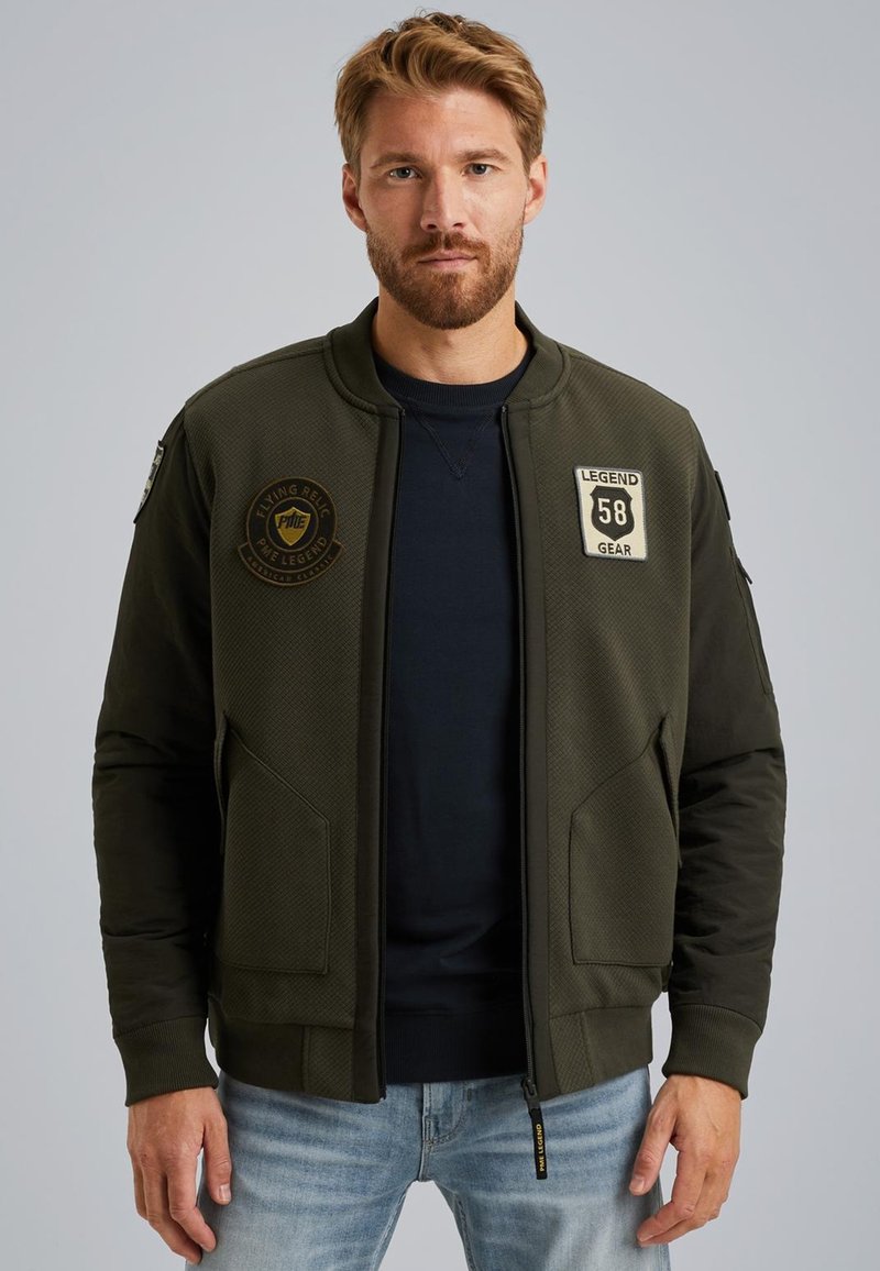 Veste bomber vert olive avec un haut texturé, fermeture éclair, poignets côtelés, deux poches avant et des patchs brodés sur la poitrine.
