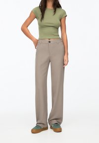 T-shirt vert ajusté, pantalon beige taille haute, et baskets vertes avec des semelles brunes. Pantalon à jambe droite avec poches avant et fermeture à bouton.