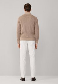 Beige geribbelde gebreide trui met een hoge kraag, gecombineerd met witte broek en donkere schoenen. Focus op textuur en een neutraal kleurenpalet.