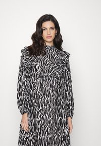 Vestido estampado preto e branco com mangas longas, detalhes em babados nos ombros, gola alta e silhueta fluida.