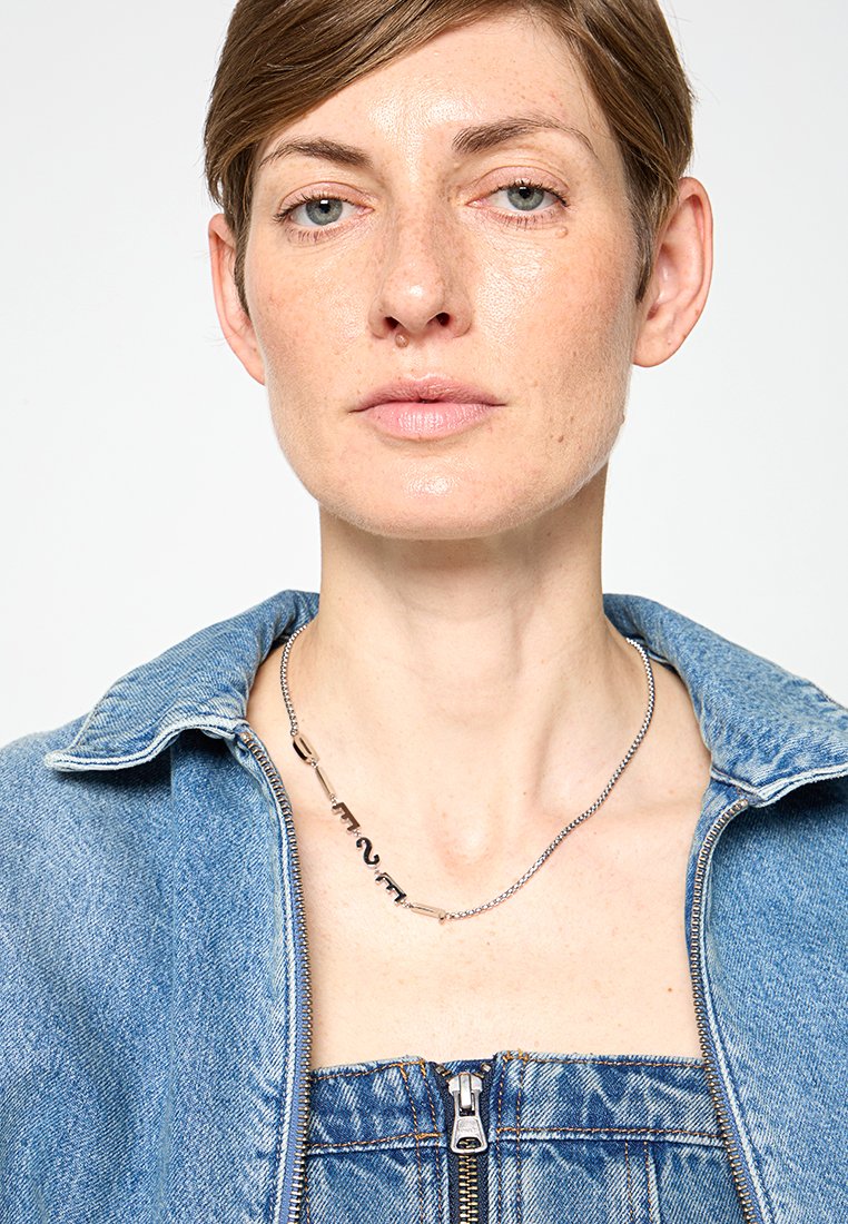 Collana in argento con maglie di forma irregolare, impreziosita da accenti di lettering audaci. Il modello indossa una giacca di jeans e una salopette.