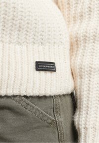 Pull en tricot crème avec un ourlet côtelé, arborant une petite étiquette noire ornée de texte blanc, associé à un pantalon texturé vert olive.