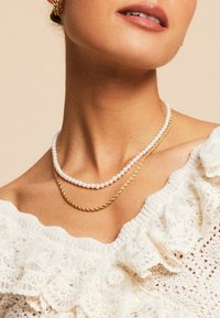 Dva slojevita ogrlicu: jedan od bijelih bisera i jedan uvrnuti zlatni lanac. Pozadina je neutralna, ističući detalje ogrlica.