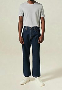 Homme portant un t-shirt gris clair à manches courtes, un jean droit bleu foncé et des baskets blanches, debout devant un fond uni.