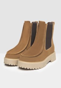 Bottines en suède marron avec des panneaux élastiques sur les côtés, des bouts ronds et des semelles en caoutchouc beige thické et texturé. Design minimaliste avec des coutures visibles.