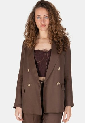 Donna con capelli lunghi e ricci che indossa un blazer doppiopetto marrone sopra un top corsetto in pizzo scuro su uno sfondo chiaro uniforme.